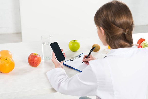 back view of dietitian holding smartphone with blank screen and writing in clipboard at table - Photo, Image
