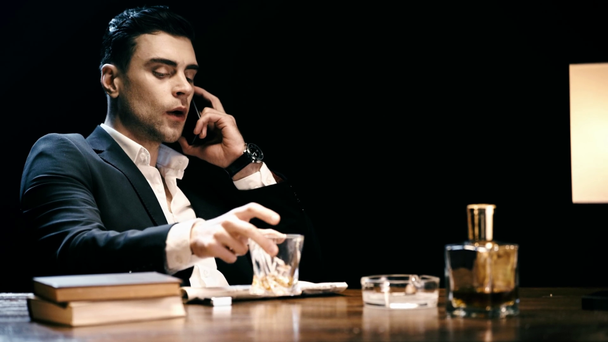 σοβαρός επιχειρηματίας κάθεται στο τραπέζι, μιλώντας για smartphone και πίνοντας ουίσκι που απομονώνεται σε μαύρο - Πλάνα, βίντεο