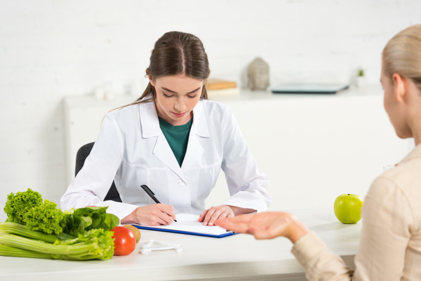 diététiste concentré en manteau blanc écrit dans le presse-papiers et patient à la table
 - Photo, image