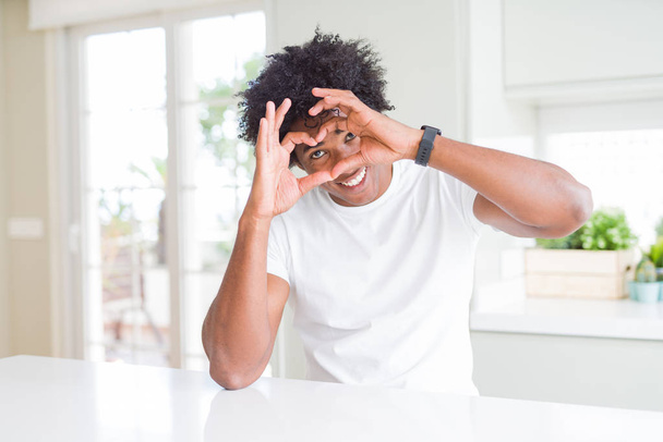 Giovane uomo afroamericano indossa casual t-shirt bianca seduta a casa Fare a forma di cuore con le mani e le dita sorridenti guardando attraverso il segno
 - Foto, immagini