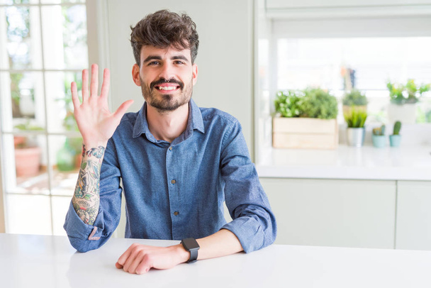 Молодий чоловік в повсякденній сорочці сидить на білому столі, показуючи і вказуючи пальцями номер п'ять, посміхаючись впевнено і щасливо
. - Фото, зображення