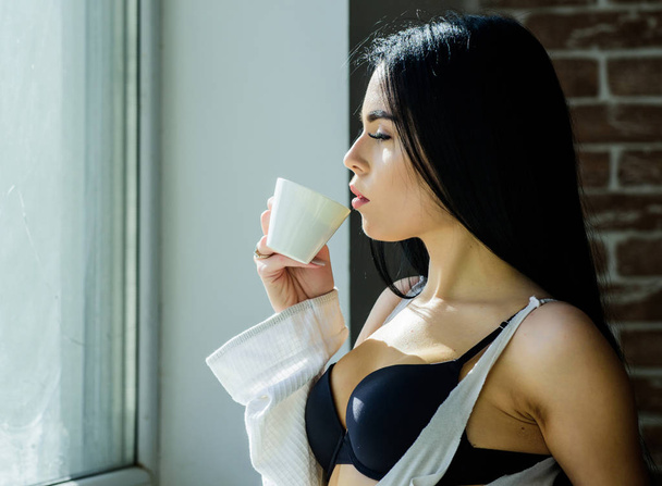Girl sexy breasts drinking coffee near windowsill. Attractive female lingerie relaxing at home. Perfect coffee time. But first coffee. Tranquility and peace. Sip of creamy cappuccino. Morning ritual - 写真・画像