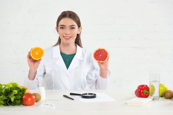 vista frontal del dietista sonriente de capa blanca sosteniendo naranja cortada y pomelo
 - Foto, Imagen
