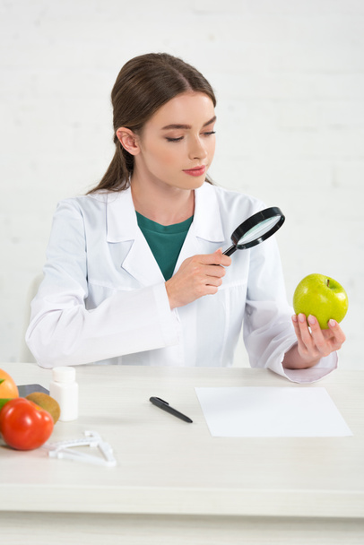 διαιτολόγος σε λευκό παλτό κοιτάζοντας μήλο μέσω μεγεθυντικού γυαλιού - Φωτογραφία, εικόνα
