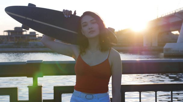 Mladá žena stojící na nábřeží při západu slunce a má na kameře skateboard. - Fotografie, Obrázek
