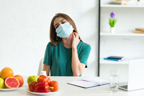 young woman in medical mask sitting at table with fruits, vegetables and pills and scratching neck at home - Foto, Imagem