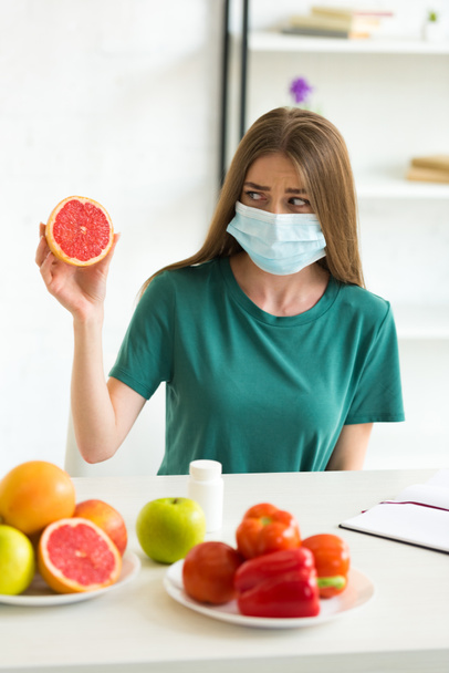 jeune femme en masque médical assis à table avec des fruits, légumes et pilules et tenant pamplemousse à la maison
 - Photo, image
