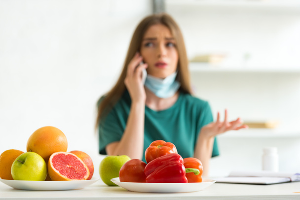 szelektív hangsúly a nő az orvosi maszkot beszél okostelefon az asztalnál gyümölcsöt, zöldséget és tablettákat otthon - Fotó, kép