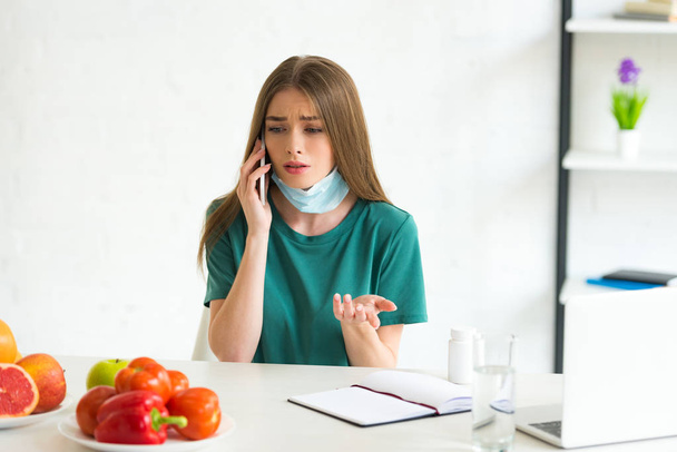 menina em máscara médica falando no smartphone à mesa com frutas, legumes e pílulas em casa
 - Foto, Imagem