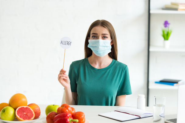 front view of woman in medical mask sitting at table with fruits, vegetables and pills and holding banner on stick with inscription allergy at home - Foto, Bild