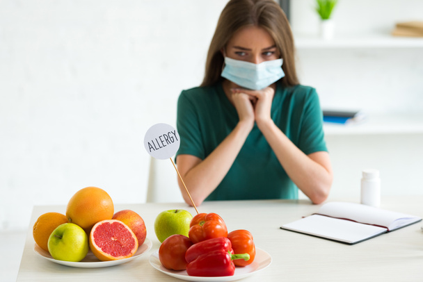 femme triste dans le masque médical appui visage avec les mains tout en étant assis à la table avec des fruits, légumes et gabarit avec l'inscription allergie
 - Photo, image