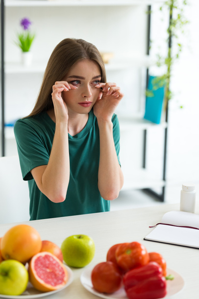 Mädchen mit Allergie wischt sich Tränen, während sie mit Obst, Gemüse und Tabletten am Tisch sitzt - Foto, Bild