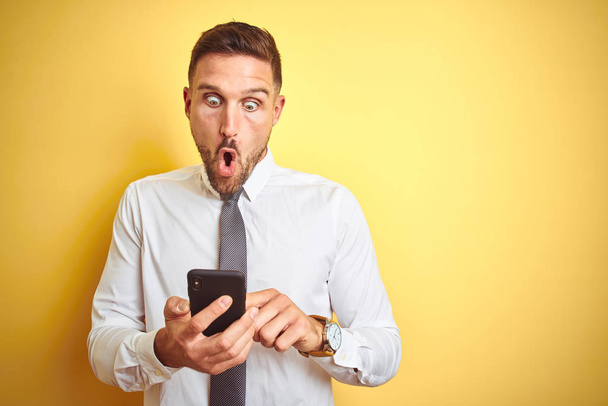 Mladý pohledný obchodní muž používající smartphone přes žlutý izolovaný podklad vyděšený v šoku s překvapením, strachem a vzrušením výrazem strachu - Fotografie, Obrázek