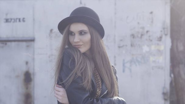 Pretty trendy girl with black stylish hat and leather jacket posing at old building in warm sunlight and hugging herself. Action. Smiling young model with smoky eyes standing outside. - Photo, image