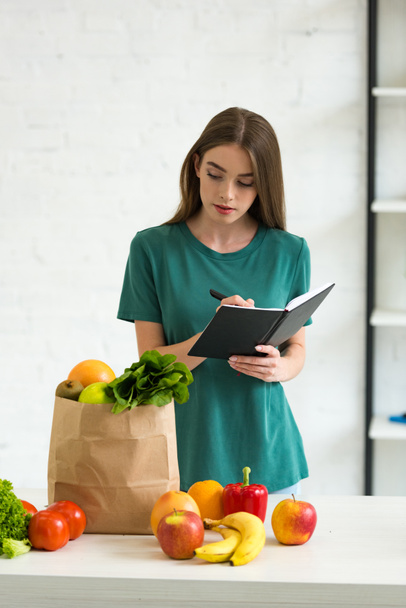 atractiva mujer sosteniendo pluma y libro de texto mientras está de pie cerca de la bolsa de papel con frutas y verduras frescas
 - Foto, imagen