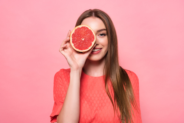 front view of attractive smiling young woman holding cut grapefruit isolated on pink - Foto, Bild