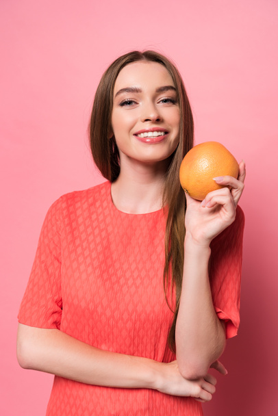attractive smiling young woman holding grapefruit and looking at camera on pink - Фото, изображение