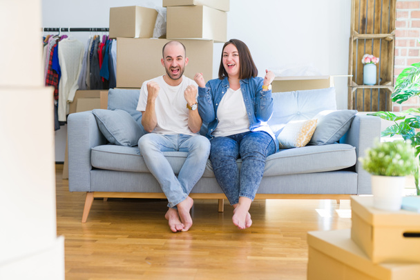Young couple sitting on the sofa arround cardboard boxes moving to a new house celebrating surprised and amazed for success with arms raised and open eyes. Winner concept. - Photo, Image