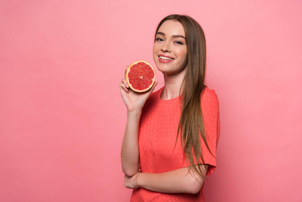 attractive smiling young woman holding cut grapefruit and looking at camera on pink - Valokuva, kuva