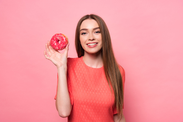 Vooraanzicht van glimlachende aantrekkelijke jonge vrouw die zoete donut vasthoudt en naar de camera op roze kijkt - Foto, afbeelding