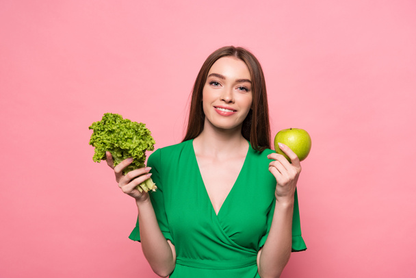 front view of attractive young woman holding lettuce and apple and looking at camera isolated on pink - Photo, Image