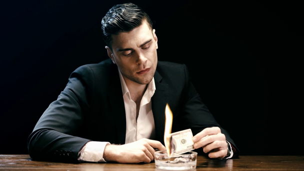 hombre de negocios sosteniendo el billete de dólar más ligero y ardiente encima del cenicero en la mesa de madera aislada en negro
 - Imágenes, Vídeo
