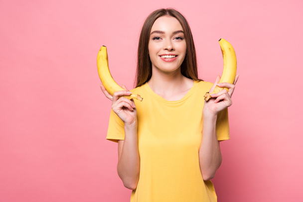 attractive smiling girl holding bananas and looking at camera on pink - Fotoğraf, Görsel