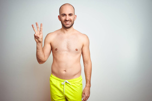 Young shirtless man on vacation wearing yellow swimwear over isolated background showing and pointing up with fingers number three while smiling confident and happy. - Photo, Image
