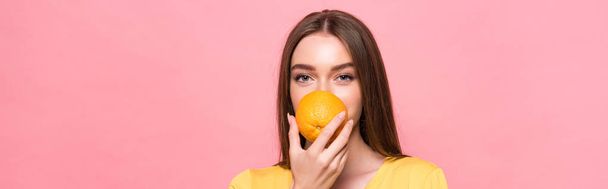 panoramic shot of young woman eating orange and looking at camera isolated on pink - 写真・画像