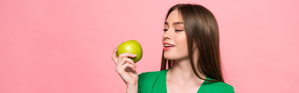 panoramic shot of attractive smiling girl holding green apple isolated on pink - Photo, Image