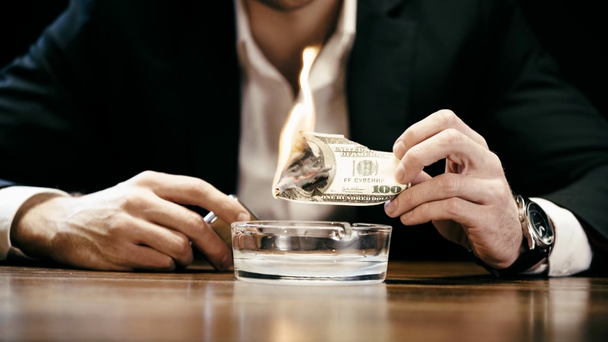περικομμένη όψη του επιχειρηματία που κρατά αναπτήρα και κάψιμο δολαρίου πάνω από τασάκι σε ξύλινο τραπέζι - Πλάνα, βίντεο