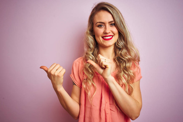 Young beautiful woman wearing t-shirt standing over pink isolated background Pointing to the back behind with hand and thumbs up, smiling confident - Photo, Image