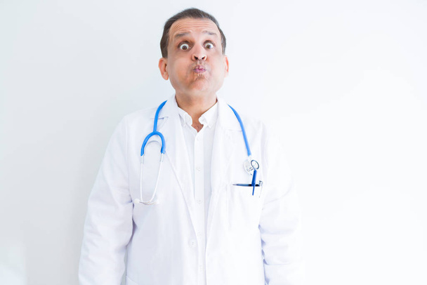 Muž středního věku doktor s stetoskem a lékařským kabátem na bílém pozadí s veselenou tváří. Ústa nafouknutými vzduchem, šíleným výrazem. - Fotografie, Obrázek