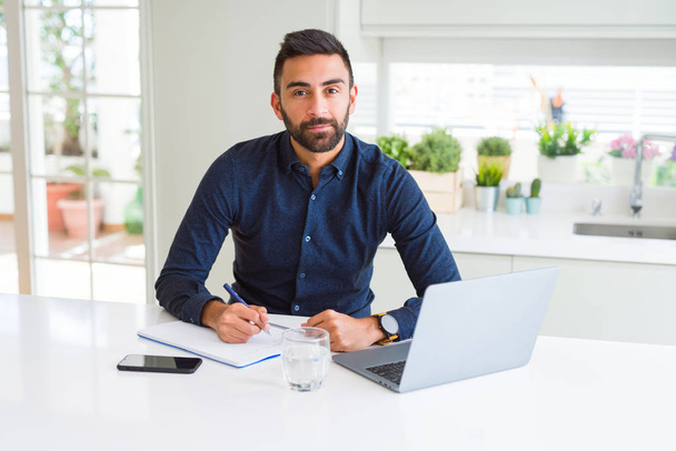 Hombre hispano guapo trabajando usando la computadora y escribiendo en un papel con una expresión de confianza en la cara inteligente pensando seriamente
 - Foto, imagen