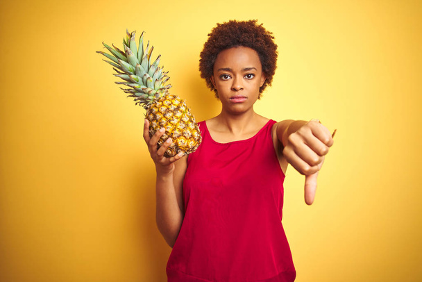 African American kobieta trzyma tropikalny ananas na żółtym białym tle z zły twarz, negatywne znak pokazując nielubię kciuki w dół, koncepcja odrzucenia - Zdjęcie, obraz