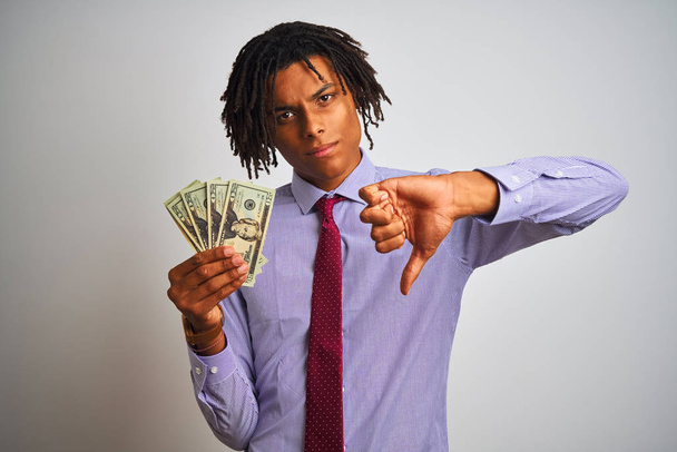 Empresário afro-americano com dreadlocks segurando dólares sobre fundo branco isolado com rosto irritado, sinal negativo mostrando antipatia com polegares para baixo, conceito de rejeição
 - Foto, Imagem