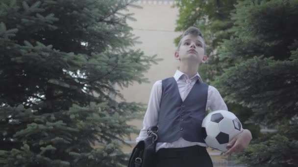 Handsome well-dressed boy standing on the street holding the soccer ball and purse. Serious young man simultaneously acting like child and adult. - Video