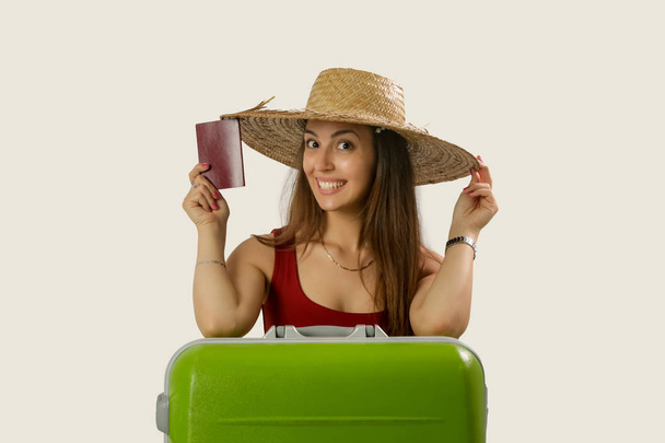 Studio portrait of a young traveler wearing a straw hat, holding passport and big green suitcase, summer vacation concept - 写真・画像