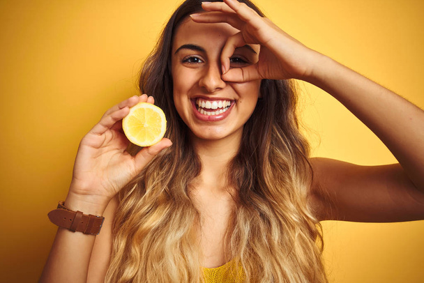 Joven hermosa mujer sosteniendo medio limón sobre fondo amarillo aislado con la cara feliz sonriendo haciendo signo bien con la mano en el ojo mirando a través de los dedos
 - Foto, Imagen