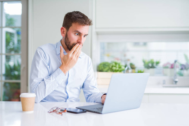 Handsome man working using computer laptop and drinking a cup of coffee cover mouth with hand shocked with shame for mistake, expression of fear, scared in silence, secret concept - Photo, image