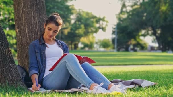 Smiling brunette woman reading magazine and writing something while sitting near the tree on grass in park - Footage, Video