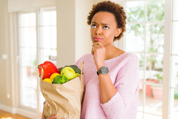 Giovane donna afroamericana in possesso di sacchetto di carta pieno di generi alimentari freschi volto serio pensando alla domanda, idea molto confusa
 - Foto, immagini