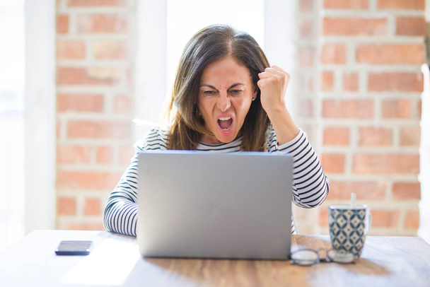 Średnim wieku starszy kobieta siedzi przy stole w domu pracy za pomocą komputera laptop zirytowany i sfrustrowani krzyczeć z gniewem, szalony i krzyczeć z podniesioną ręką, koncepcja gniew - Zdjęcie, obraz