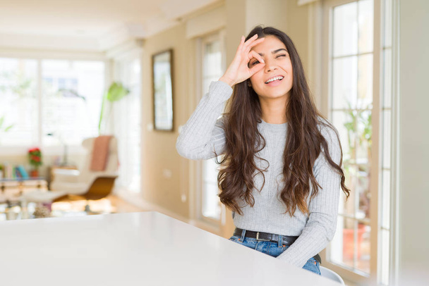 Young beautiful woman at home doing ok gesture with hand smiling, eye looking through fingers with happy face. - Photo, Image