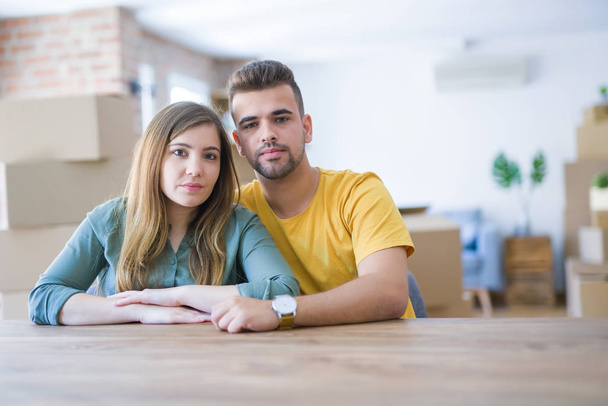 Young couple sitting on the table movinto to new home with carboard boxes behind them with serious expression on face. Simple and natural looking at the camera. - Photo, Image