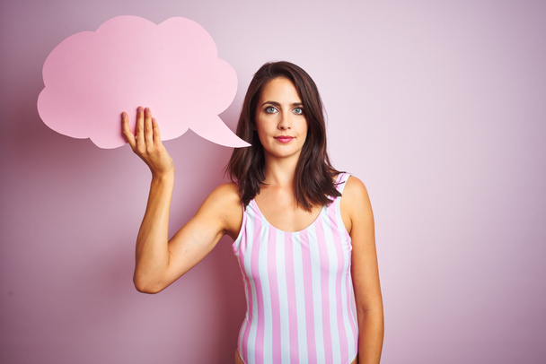 Beautiful woman wearing striped pink swimsuit and holding talking balloon over pink background with a confident expression on smart face thinking serious - Photo, image