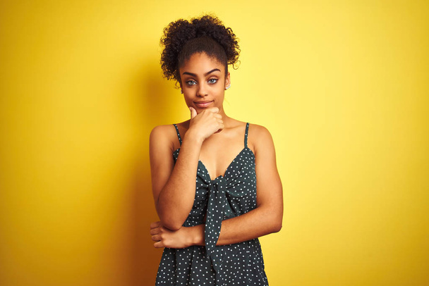 African american woman wearing summer casual green dress over isolated yellow background looking confident at the camera with smile with crossed arms and hand raised on chin. Thinking positive. - Фото, зображення