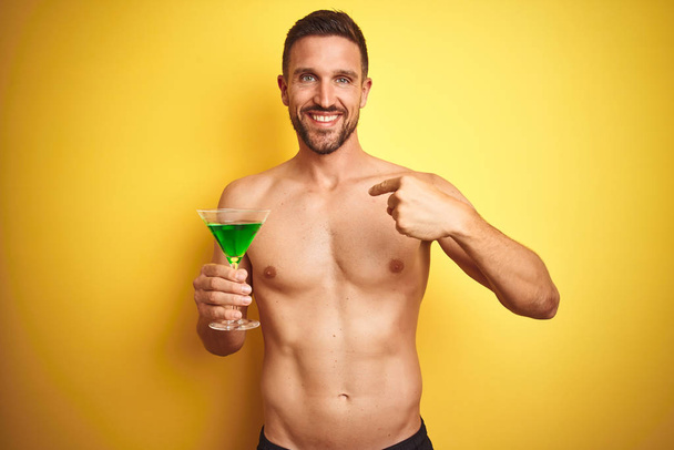 Mladý pohledný muž, který pije Letní koktejl přes izolované žluté pozadí s překvapením, ukazujícími prstem na sebe - Fotografie, Obrázek