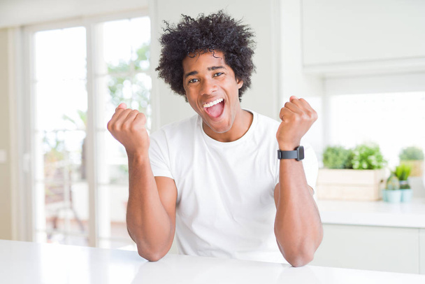 Jonge Afro-Amerikaanse man dragen casual wit t-shirt zittend thuis erg blij en opgewonden doen Winner gebaar met armen opgevoed, glimlachend en schreeuwen voor succes. Viering concept. - Foto, afbeelding
