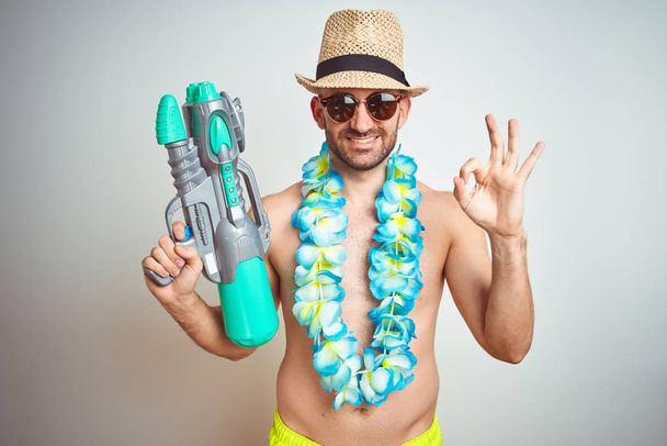 Jonge man dragen zomer hoed en Hawaiian lei bloemen Holding water gun over geïsoleerde achtergrond doen OK teken met de vingers, uitstekende symbool - Foto, afbeelding